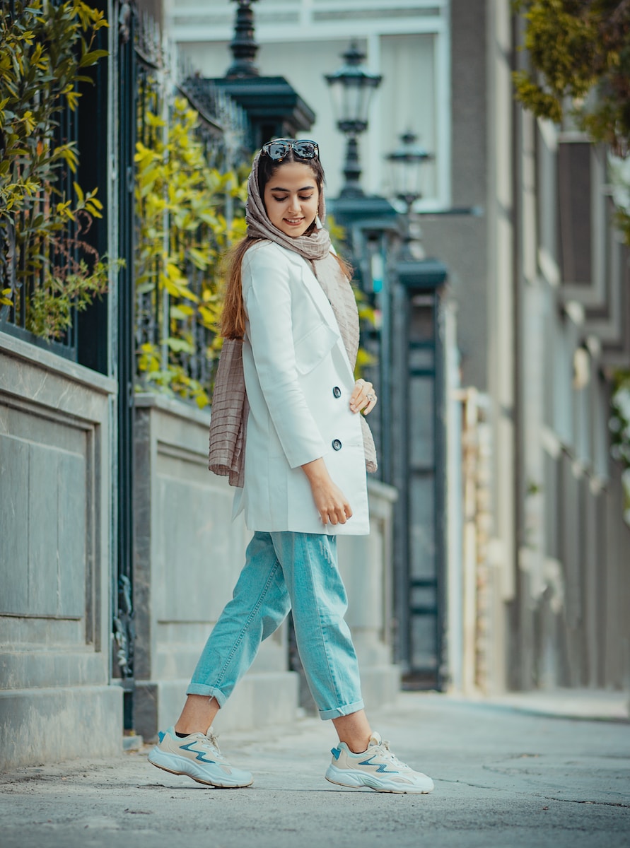 Photo by Elyas Pasban woman in white coat and blue denim jeans standing near white concrete building during daytime